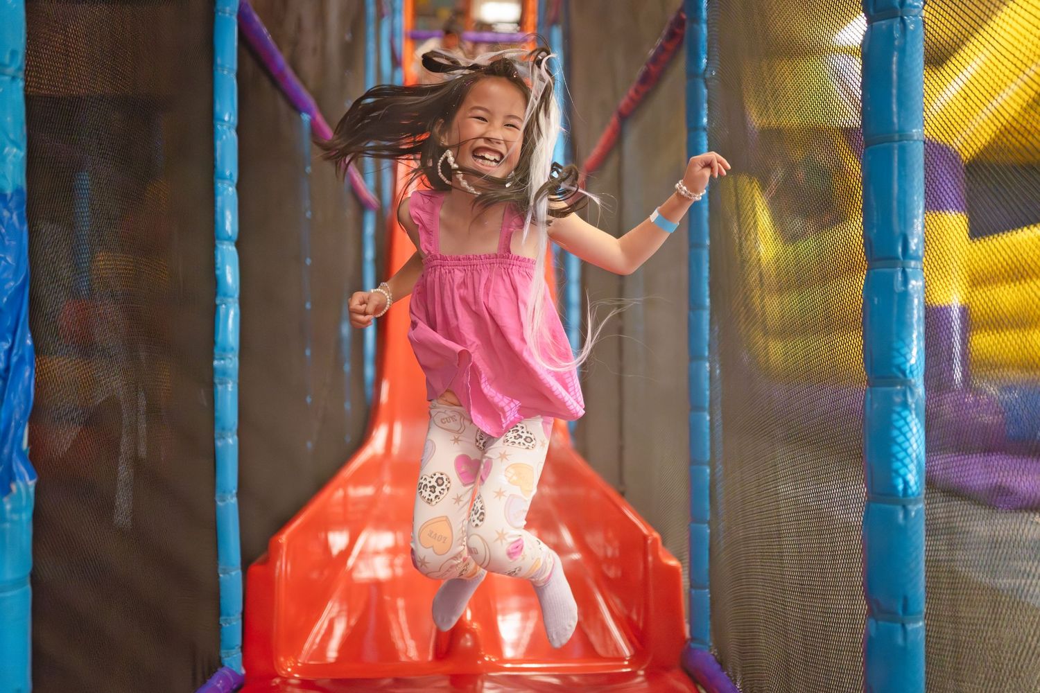 Happy girl jumping on colorful indoor playground trampoline, wearing pink dress and leggings, laughing with joy.