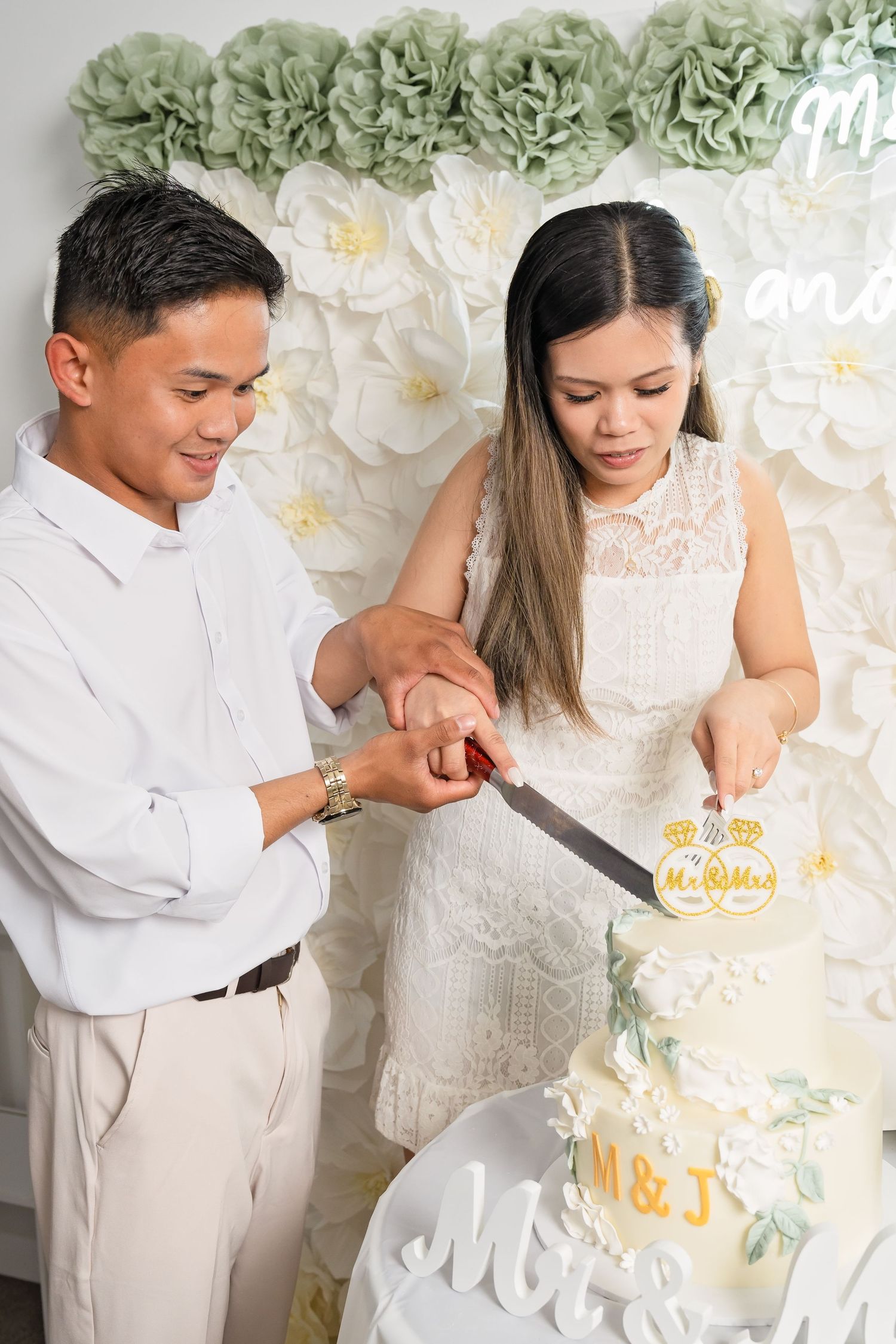 Couple cutting their wedding cake together in front of a floral backdrop, dressed in white wedding attire.