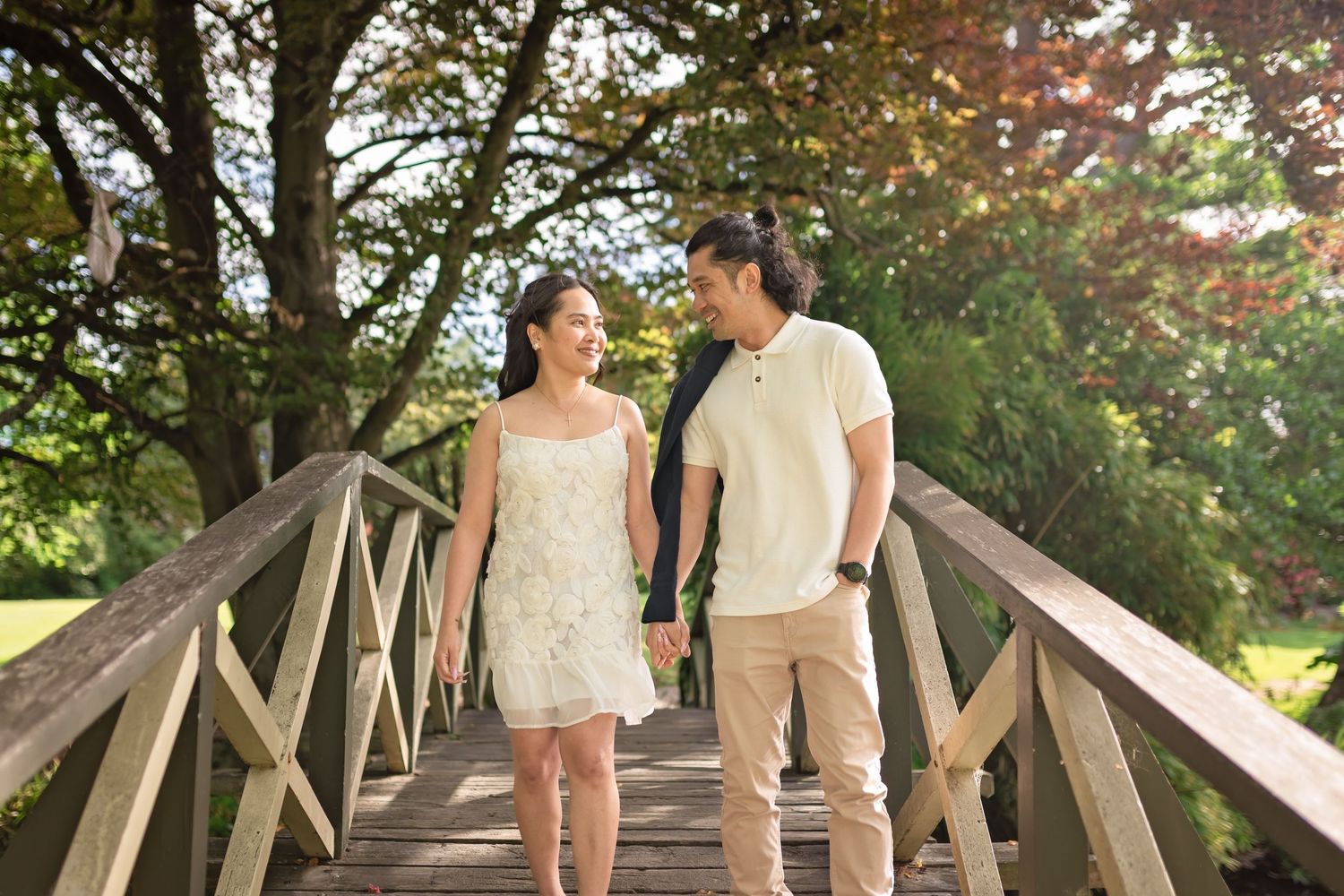 Happy couple holding hands while walking across a wooden bridge in a sunny park setting.