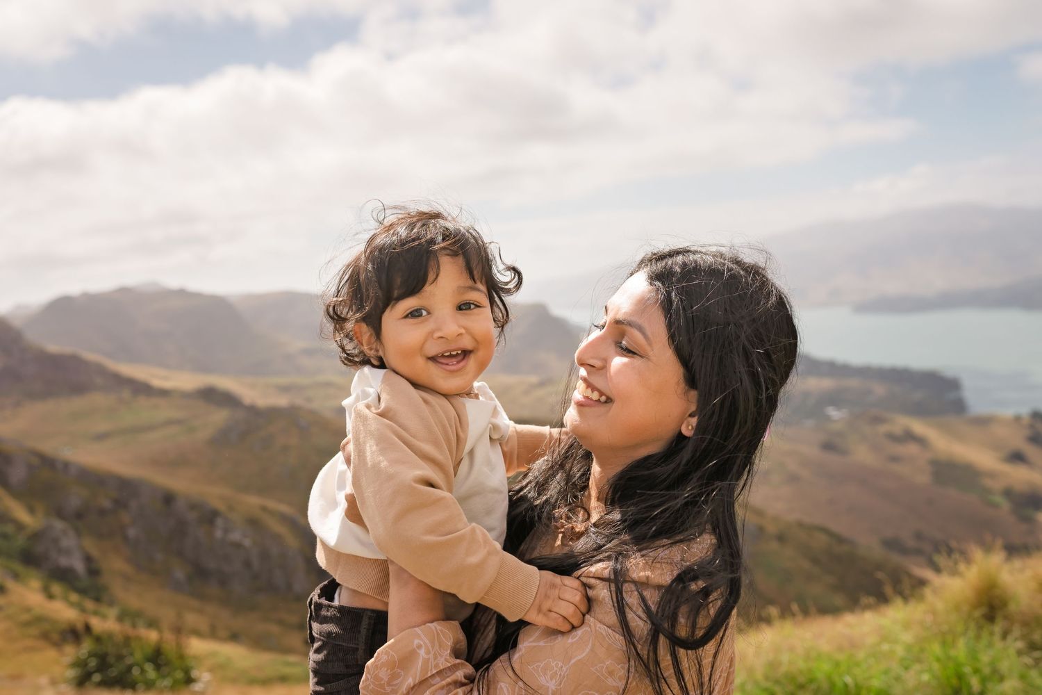 A joyful mother holds her laughing toddler against a scenic mountain landscape under a cloudy sky.