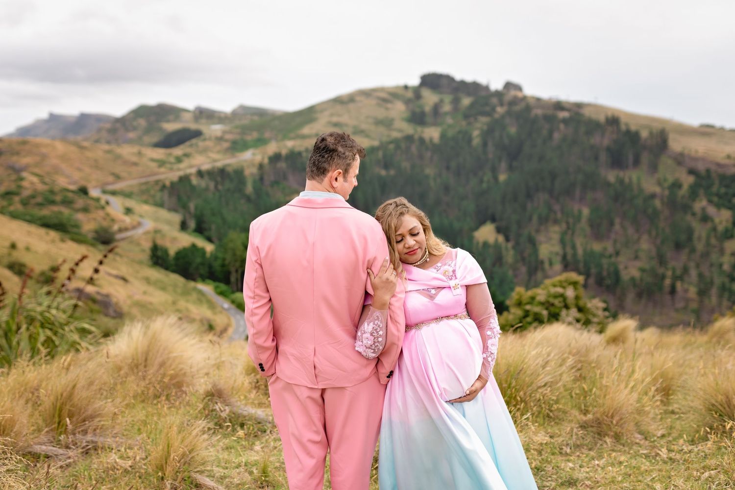 Pregnant couple in pink suit and ombre maternity gown posing on scenic mountain hillside.