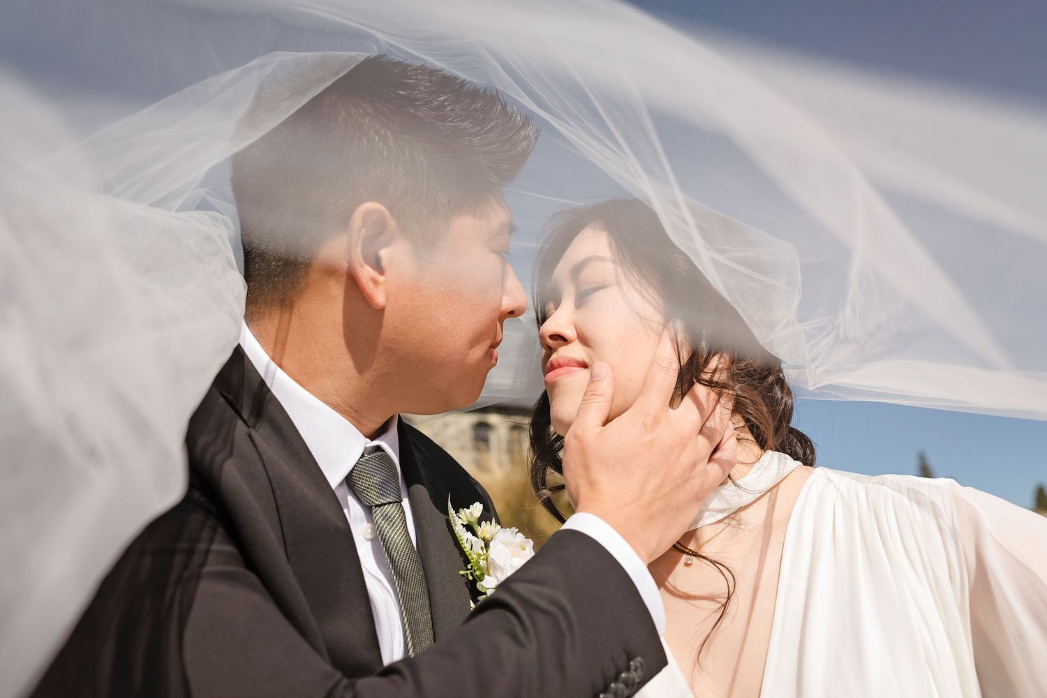 Bride and groom share a tender moment under flowing white veil with groom gently cupping brides face outdoors.