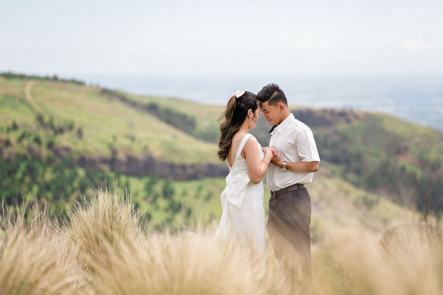 Couple sharing a romantic moment in golden grass fields with lush green hills backdrop on their wedding day.