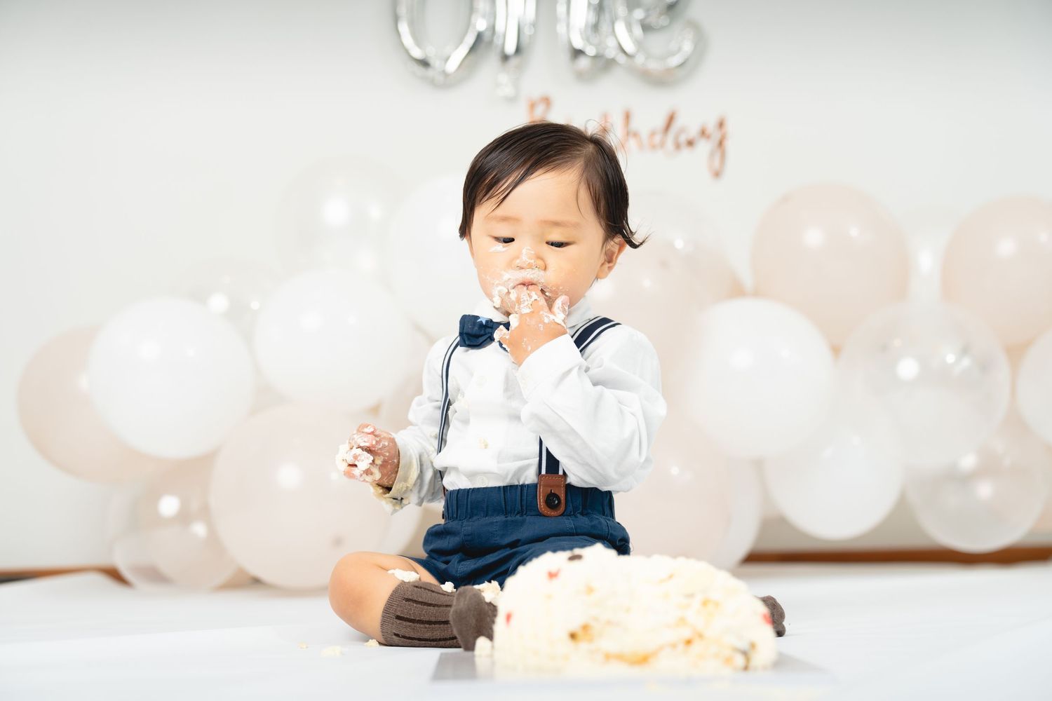 Baby in overalls enjoying birthday cake with balloons and decorations.