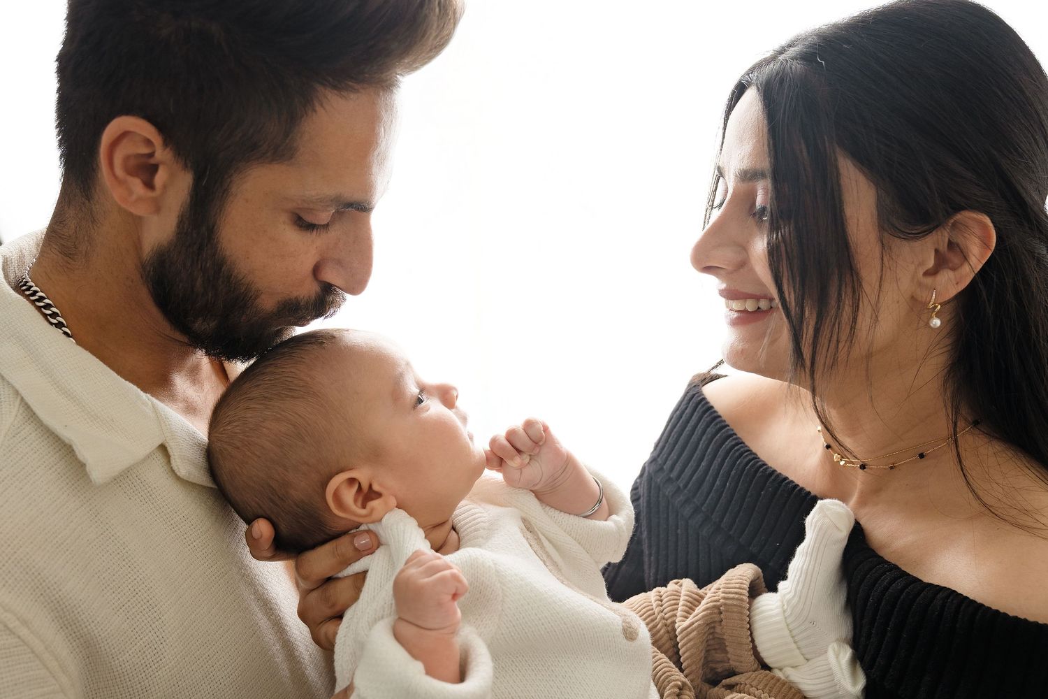 A young family shares a tender moment together with their newborn baby dressed in white.