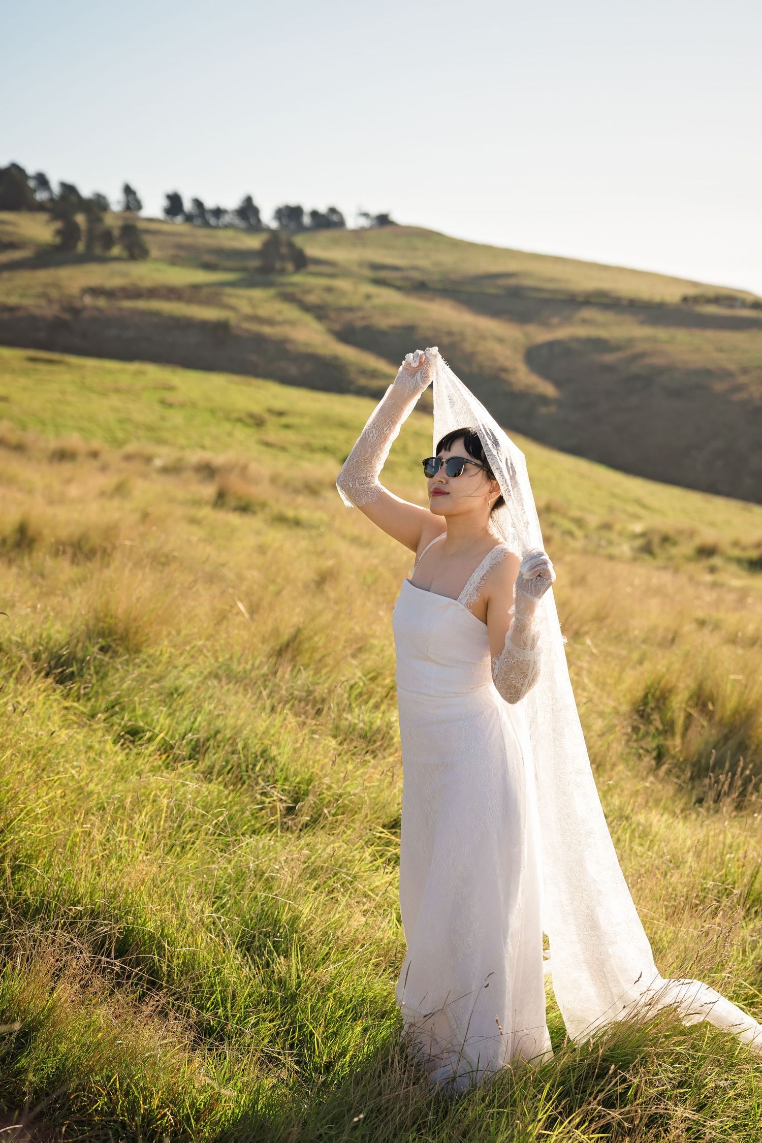 Bride in white gown and sunglasses holds veil overhead in sunny green hillside meadow.