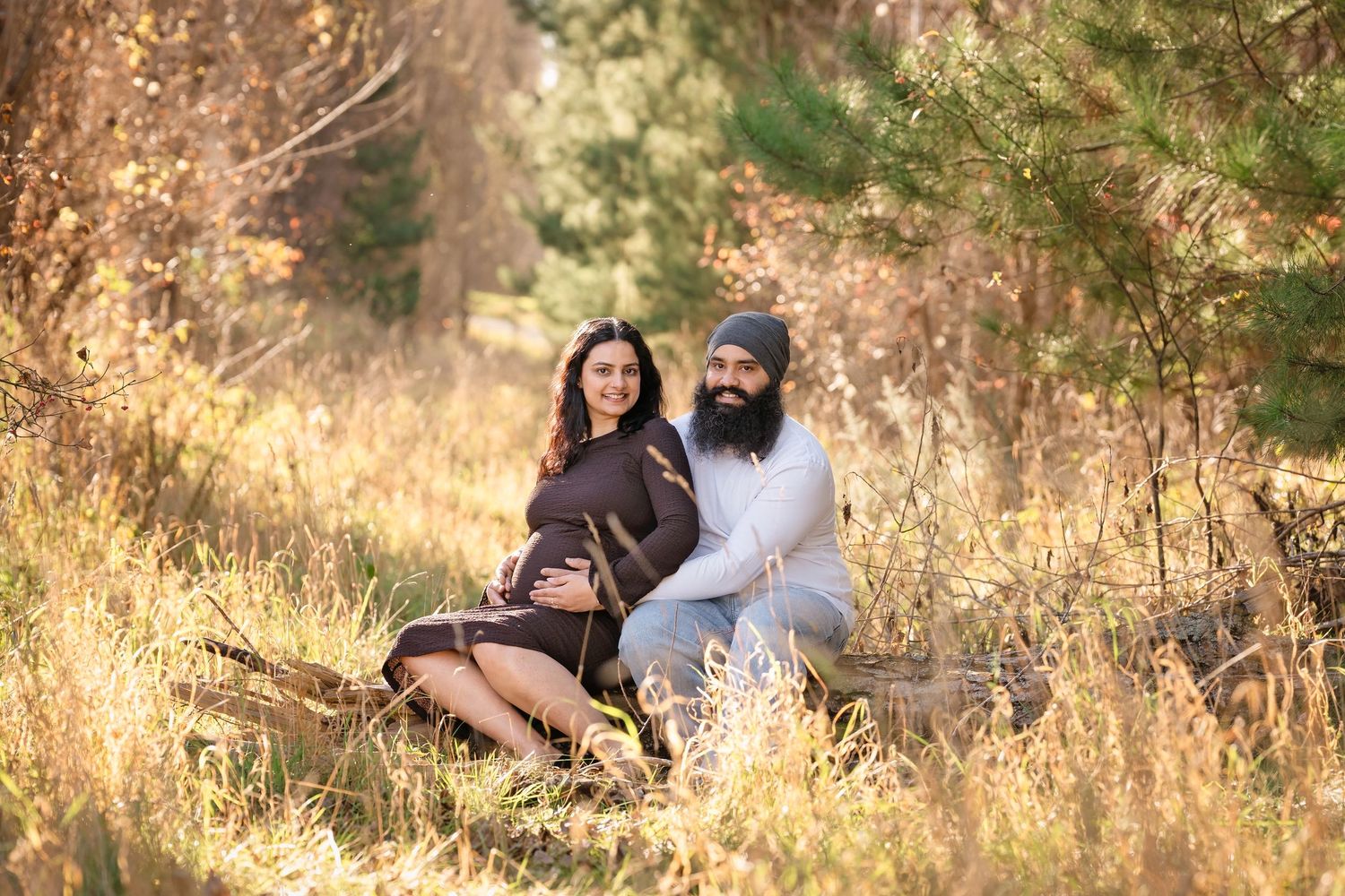 Couple sitting in golden autumn field, pregnant woman in brown dress with bearded man in gray, surrounded by tall grass and trees.
