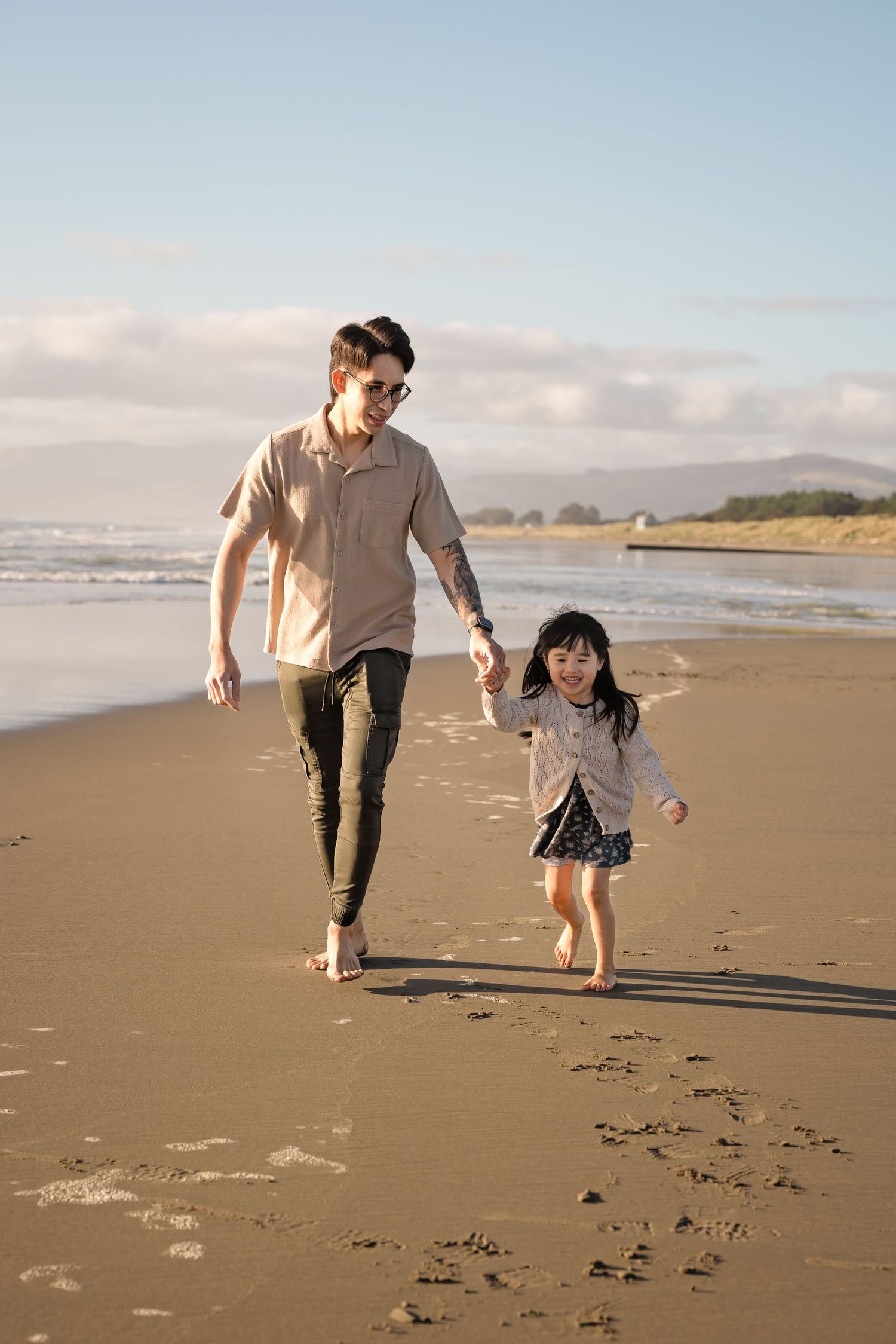 Father and young daughter joyfully running hand-in-hand along a sunny beach shoreline together.