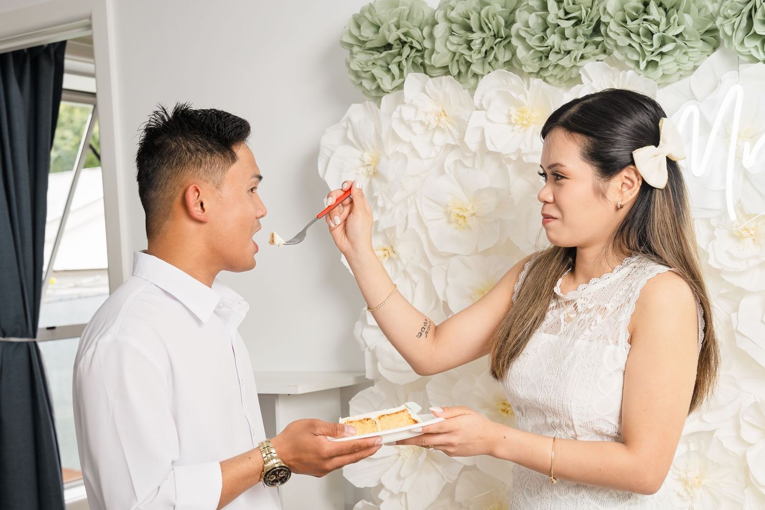 Couple in white attire feeding cake to each other at a wedding celebration with floral backdrop.