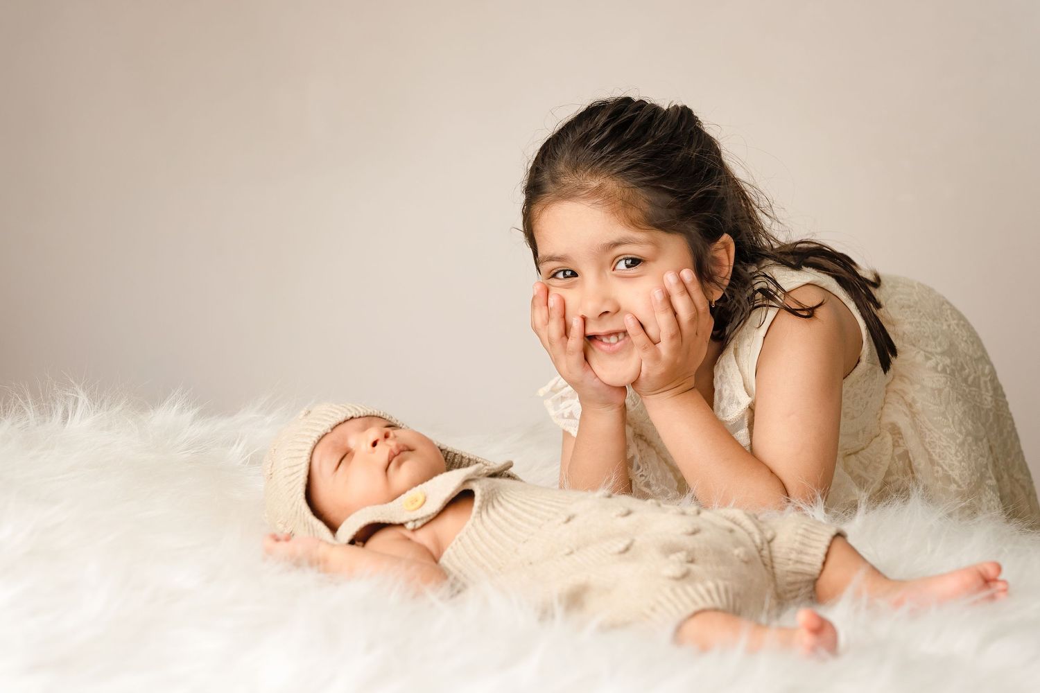 Young girl poses cheek-in-hands beside sleeping newborn baby on white fluffy blanket in studio photo.