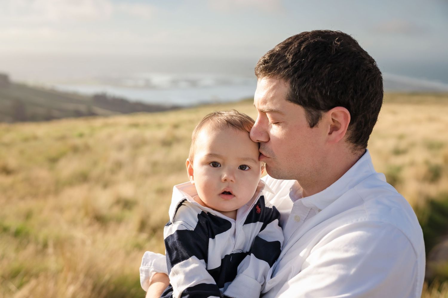 Father kissing baby outdoors in grassy coastal field, infant in striped outfit looking at camera.