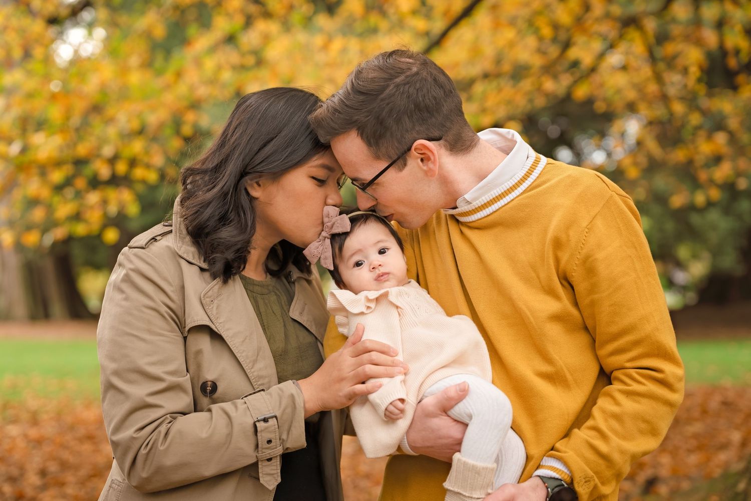 A couple leans in to kiss their smiling baby outdoors among autumn yellow foliage in a park.