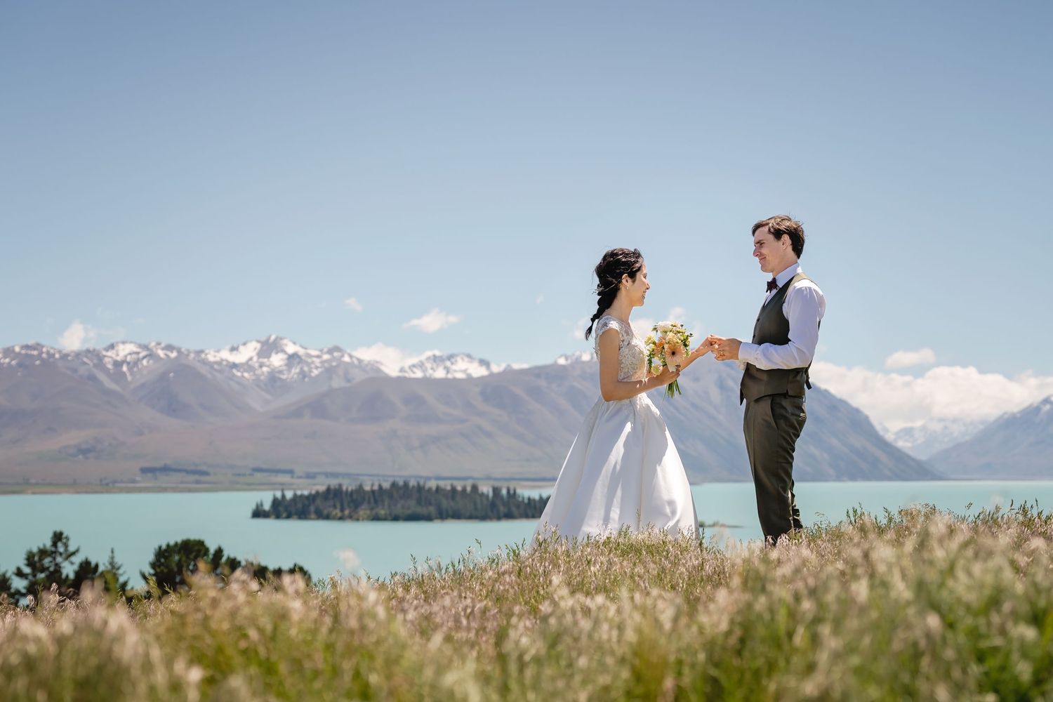 Bride and groom exchanging vows in field overlooking stunning turquoise lake and snowy mountains.