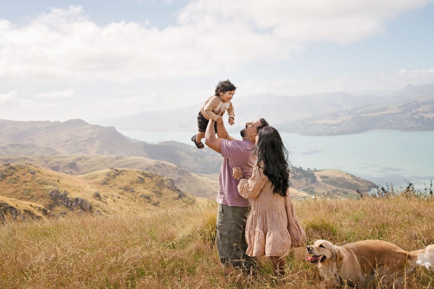 Family with baby, dog on scenic hilltop overlooking blue bay and mountains on a sunny day.
