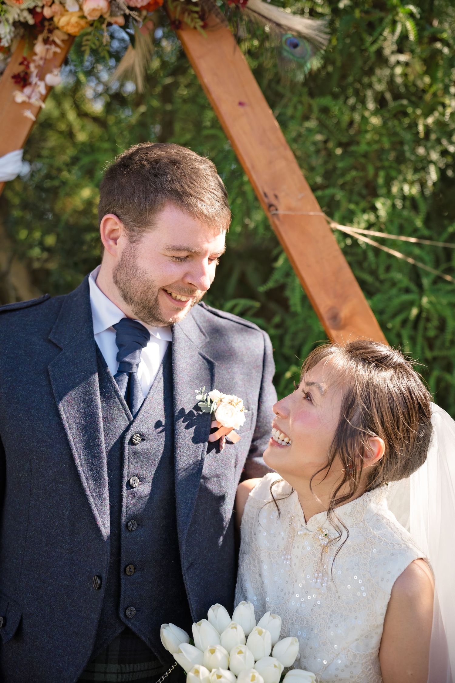 Laughing bride and groom share joyful moment under decorated wooden arch at outdoor wedding.