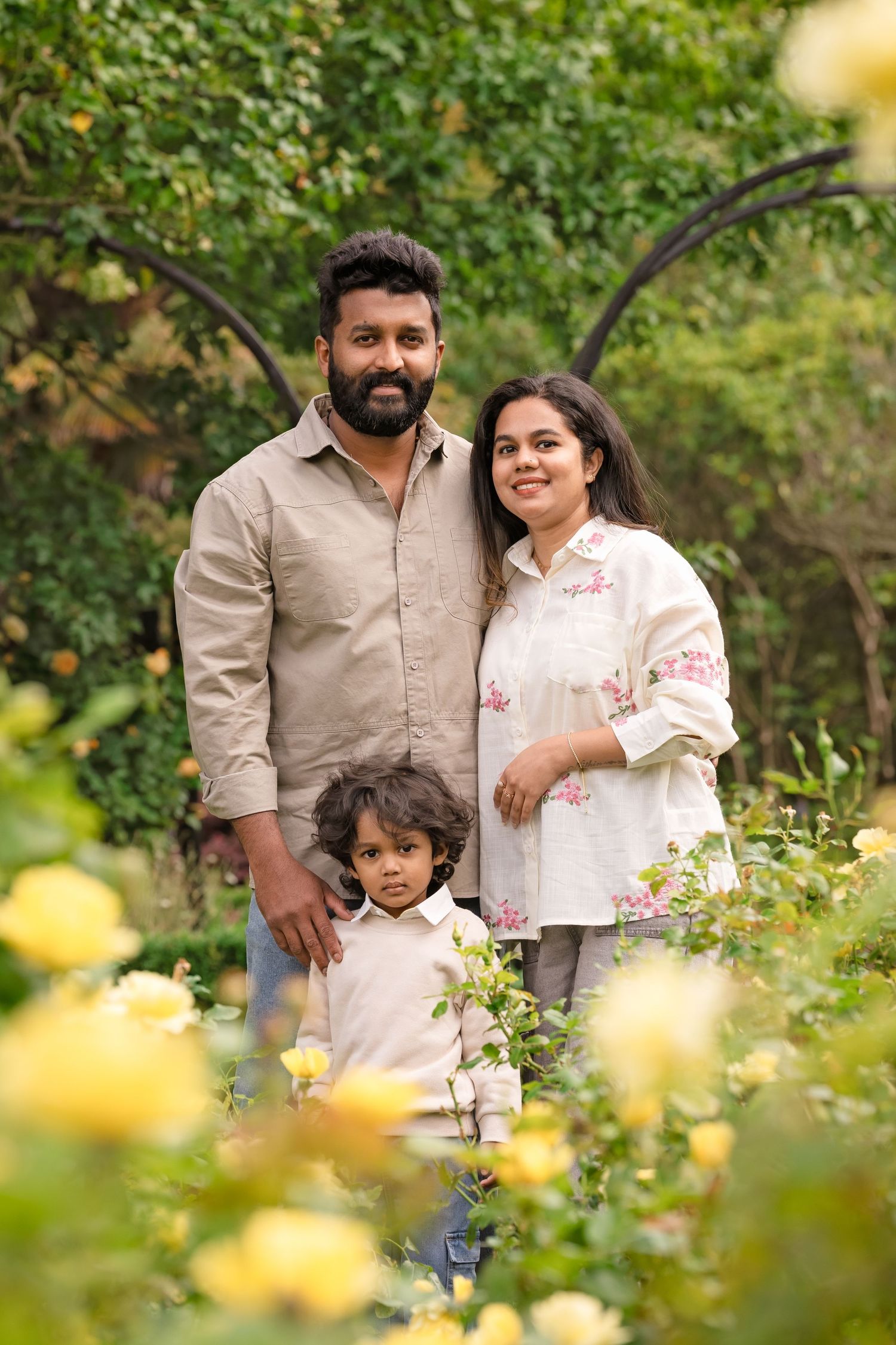 A happy family of three poses among yellow roses in a lush garden, parents smiling with their young child.
