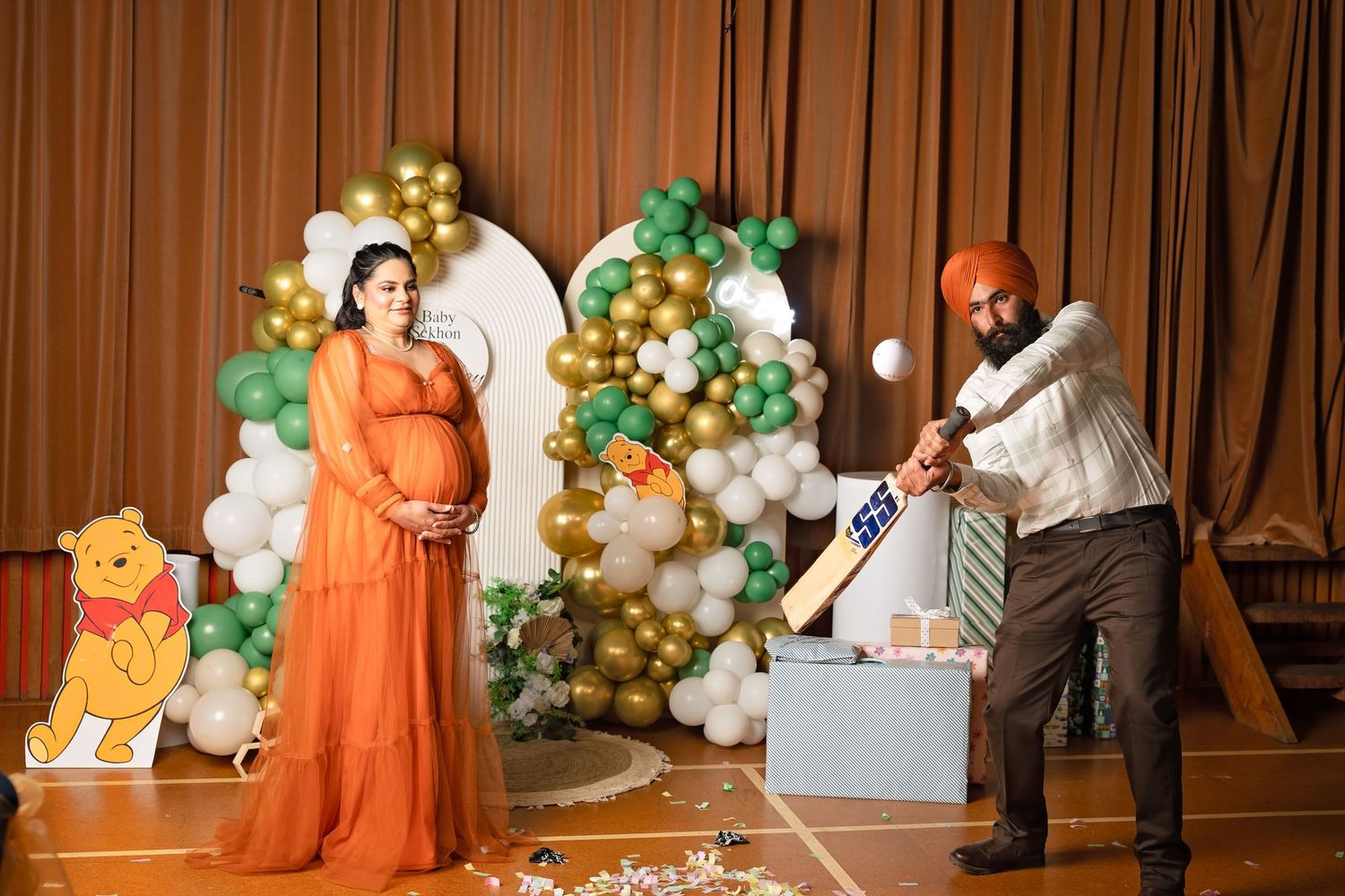 Pregnant woman in orange gown at Winnie the Pooh baby shower while man swings bat at gender reveal box.