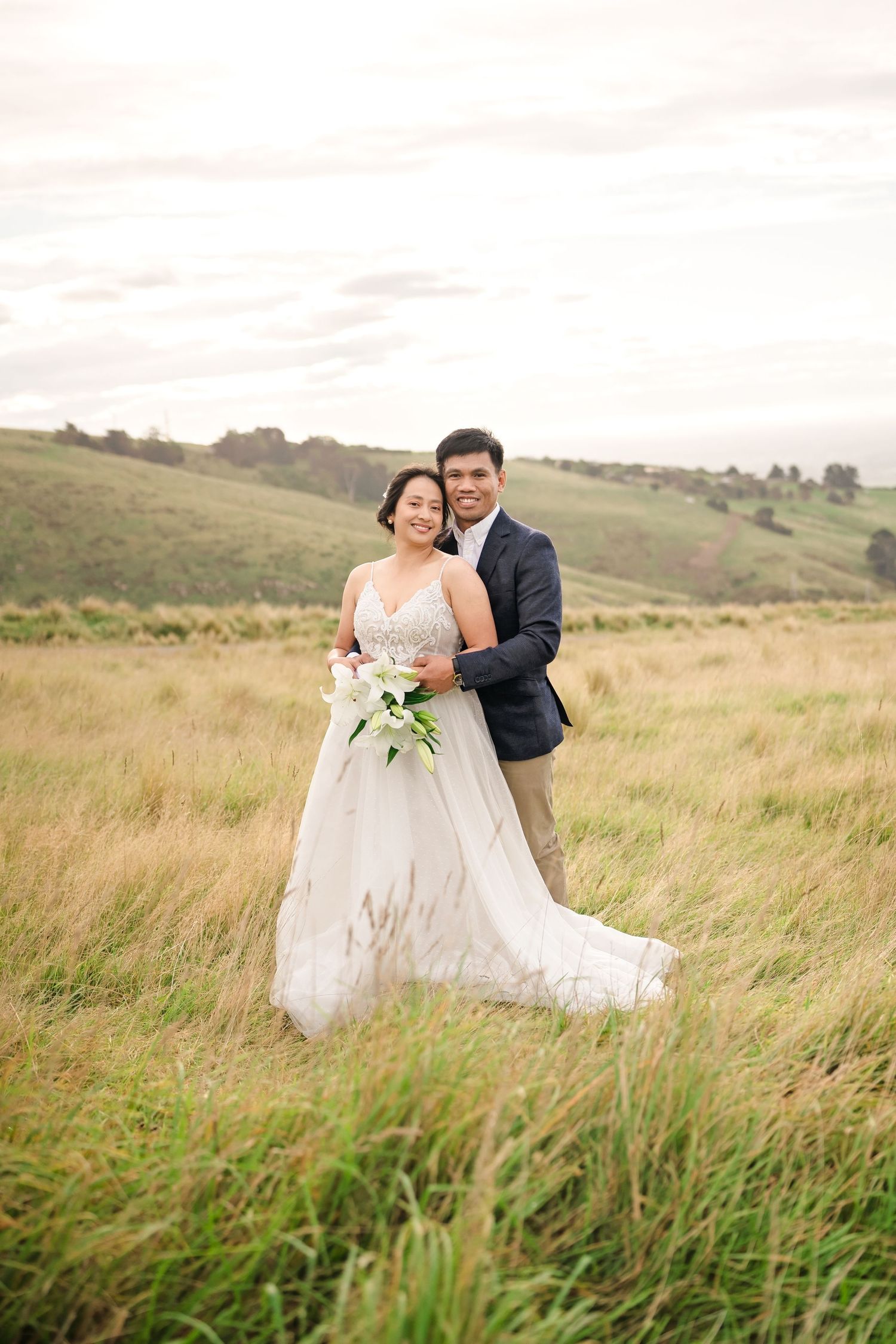 Bride and groom embracing in a lush green field, bride holding white bouquet, wearing elegant gown, groom in navy suit.