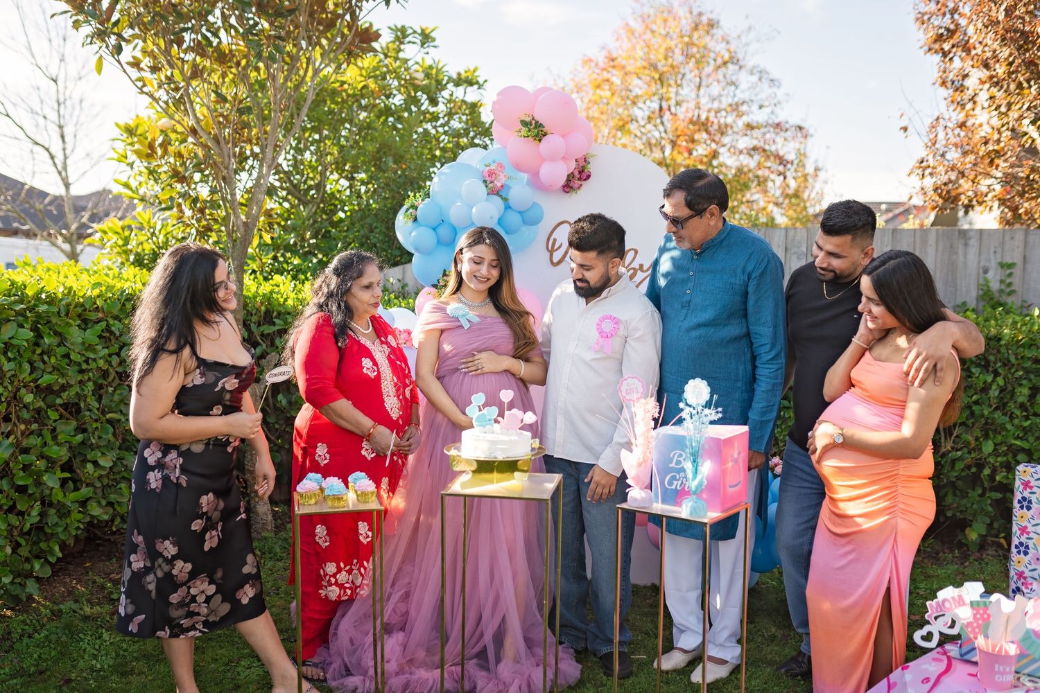 Gender reveal party celebration in a garden with pink and blue balloons and decorated gift boxes.