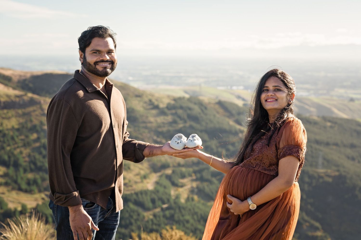 Pregnant couple holding tiny baby shoes on a scenic mountain overlook during a maternity photoshoot.