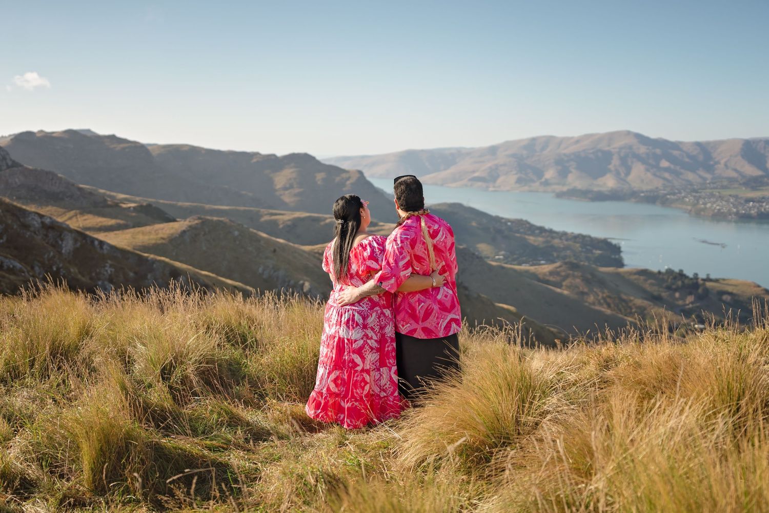 Couple in vibrant pink outfits embracing on a hilltop overlooking a scenic lake and mountain landscape.