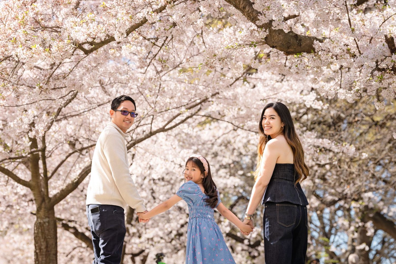 Family of three holding hands under blooming cherry blossom trees in spring, smiling and looking back at camera.