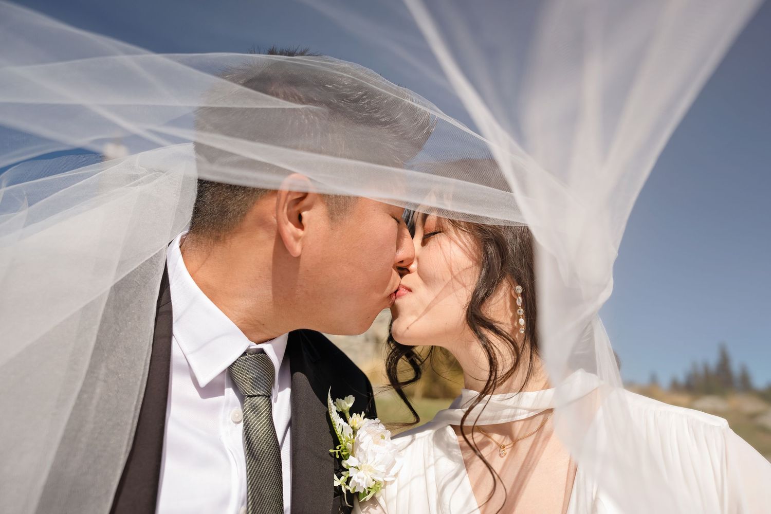 Bride and groom share a romantic kiss under a flowing white wedding veil against a bright blue sky.