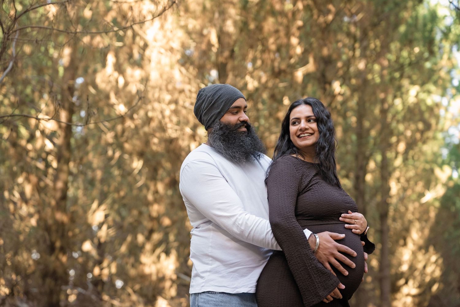 Sikh couple maternity photo shoot outdoors, man holding pregnant woman's belly, surrounded by golden trees.