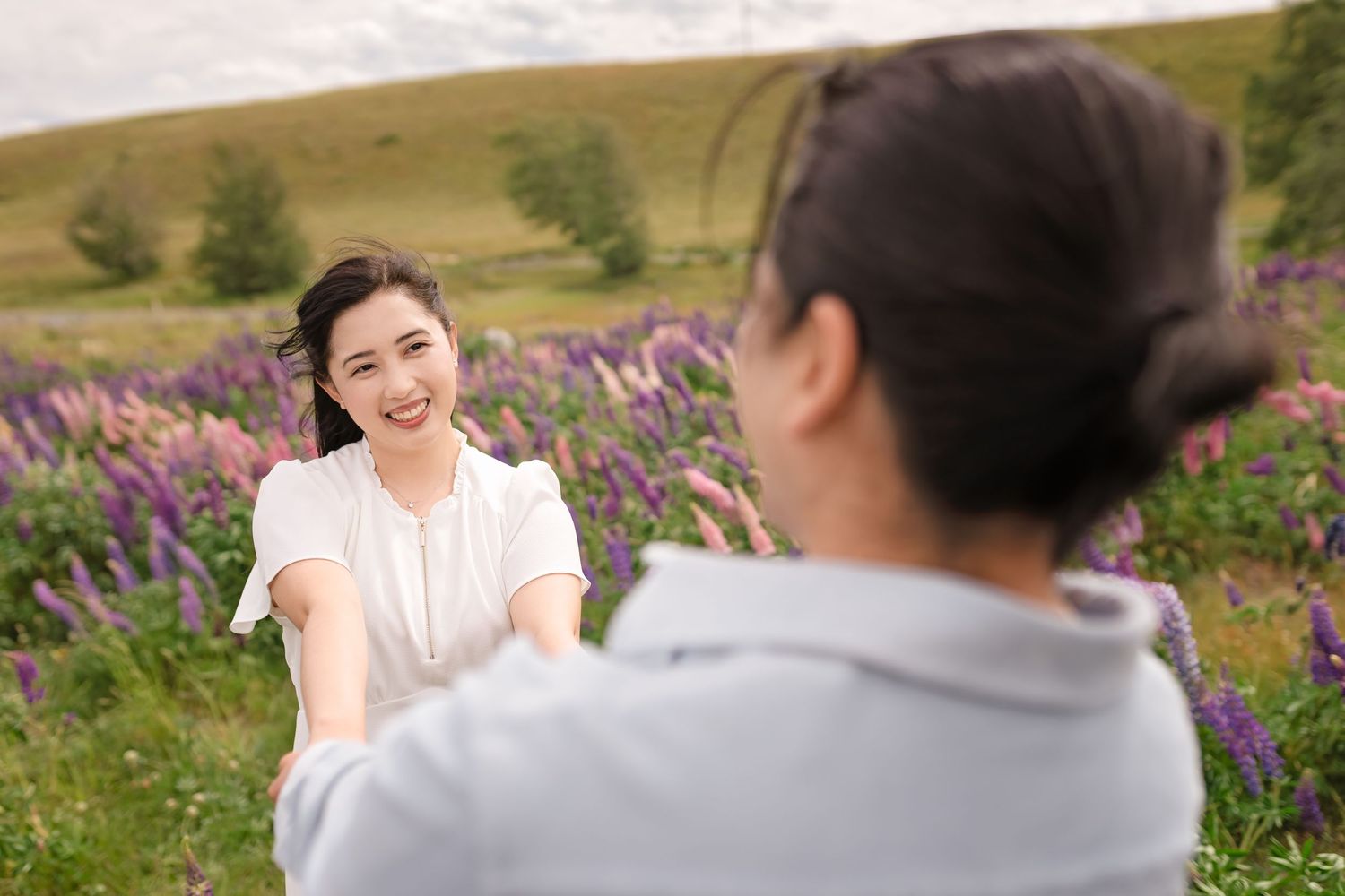 A smiling Asian woman in a white top reaches out her hand in a vibrant purple lupine flower field.