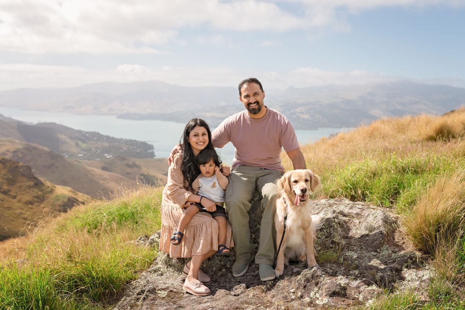 Family with baby and golden retriever dog posing on rocky hilltop with scenic lake and mountains backdrop.