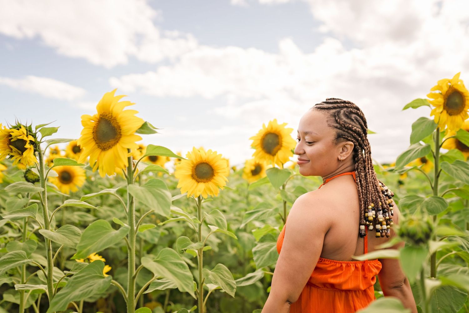 Woman in orange dress smiling among tall sunflowers in a field under a cloudy sky.