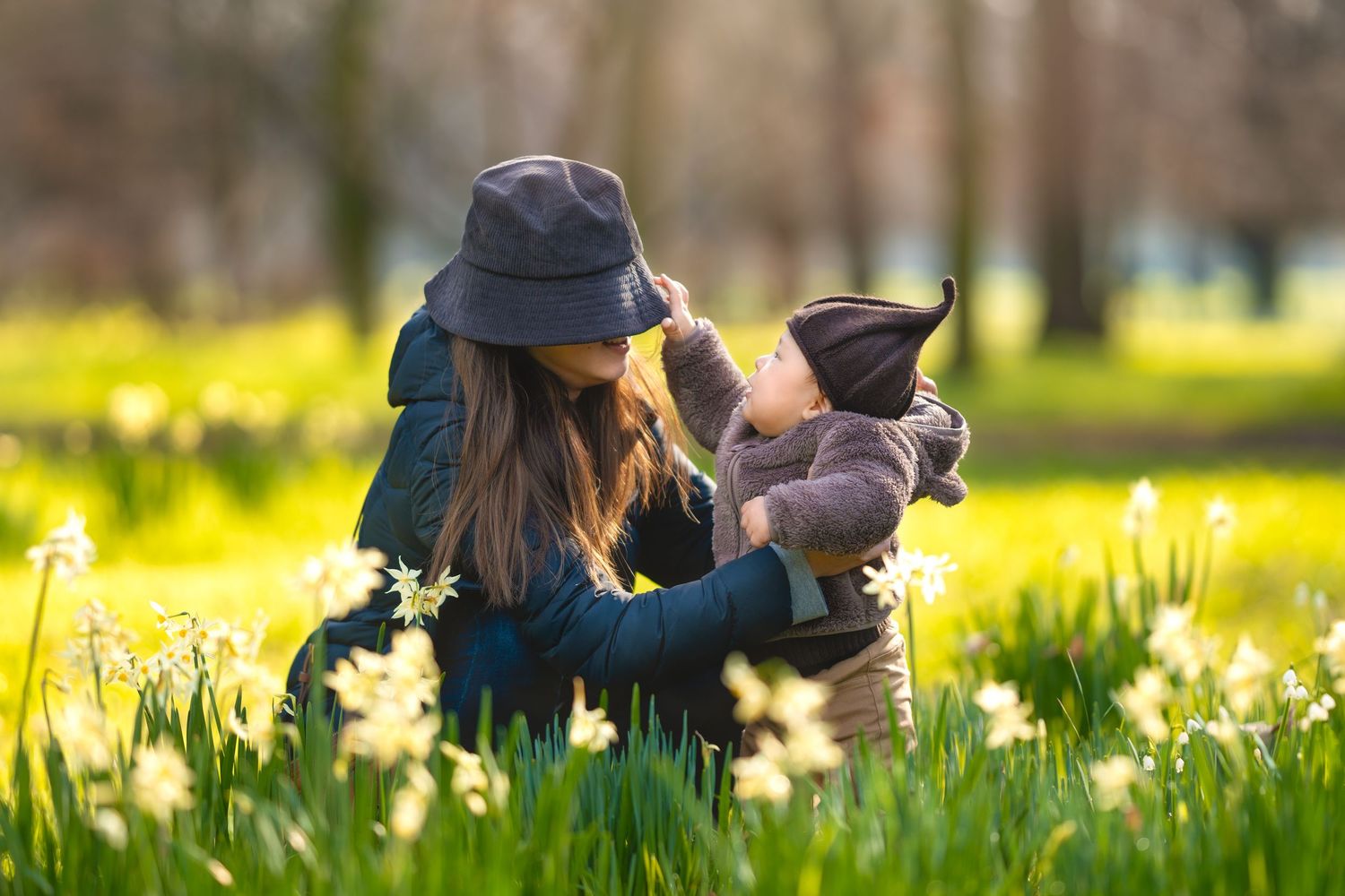 Mother holding baby in sunny spring park with blooming daffodils and lush green trees.