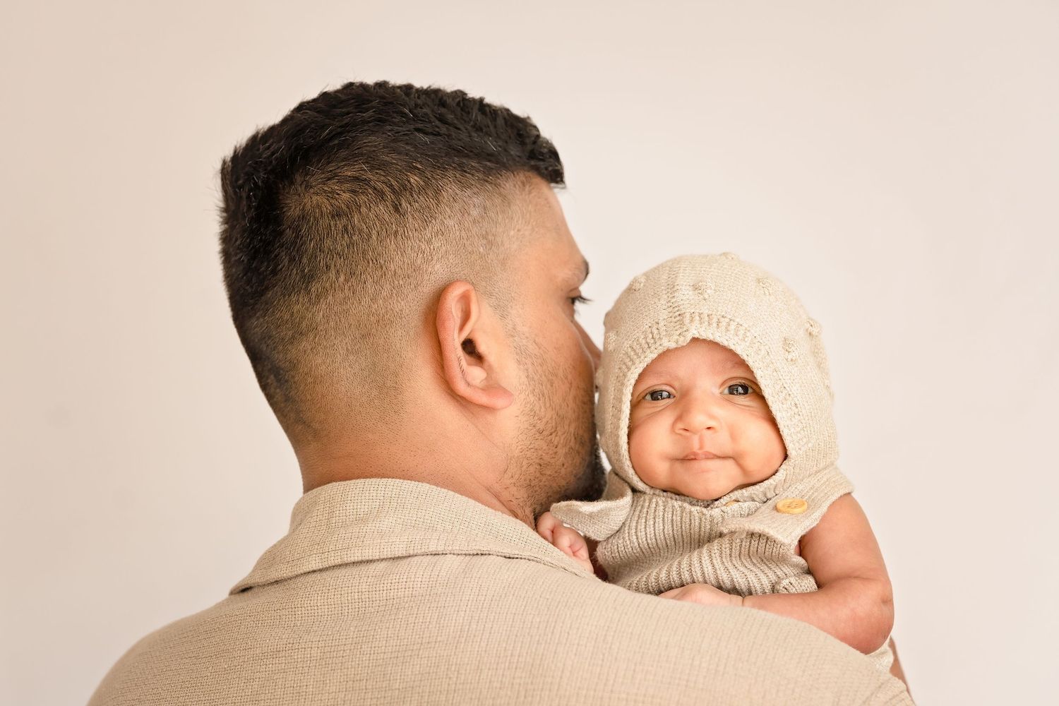 A father holds his smiling baby wearing a knit bonnet hat against a neutral background.