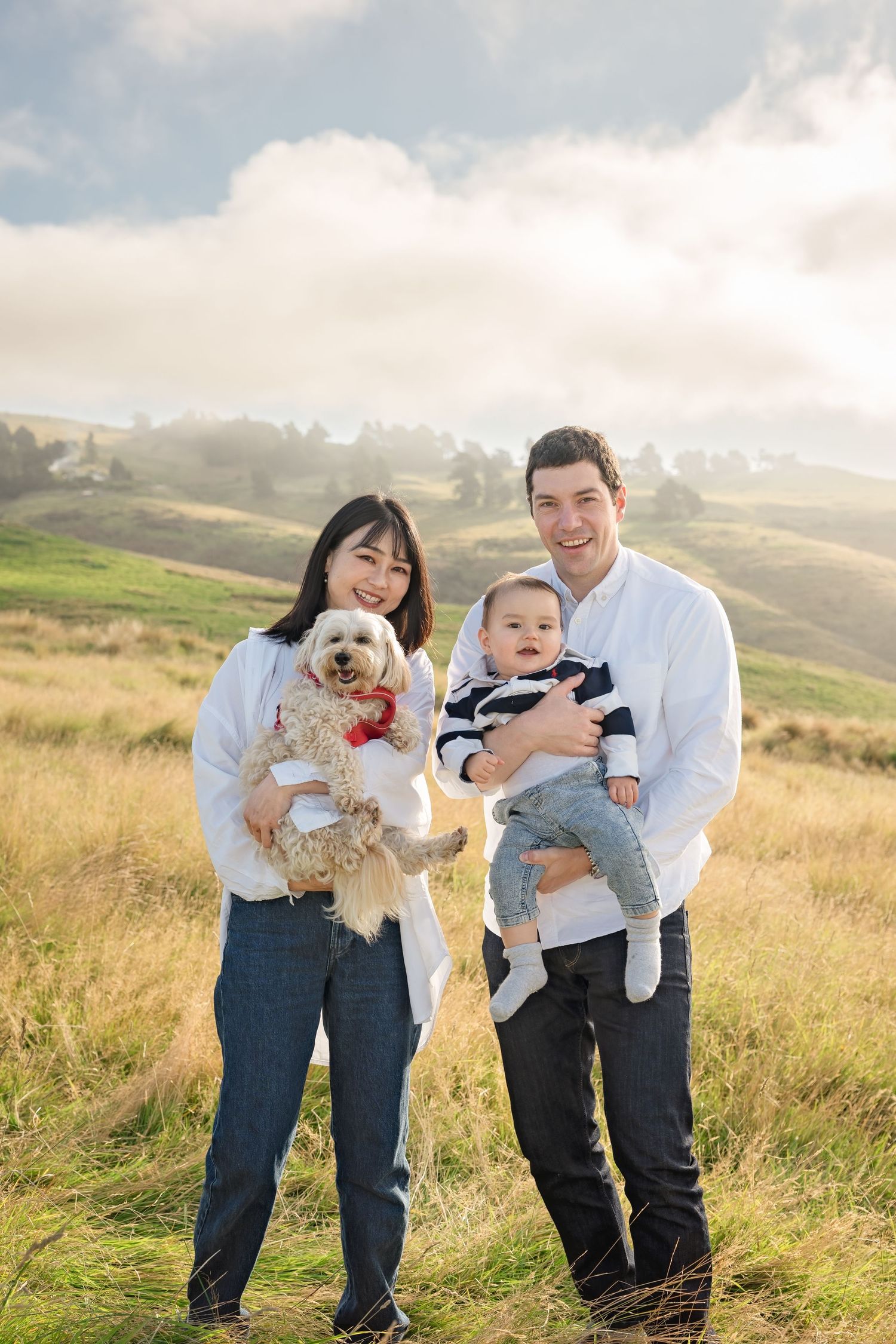 Young family posing outdoors in golden field, woman holding fluffy dog, man carrying baby, misty hills background.