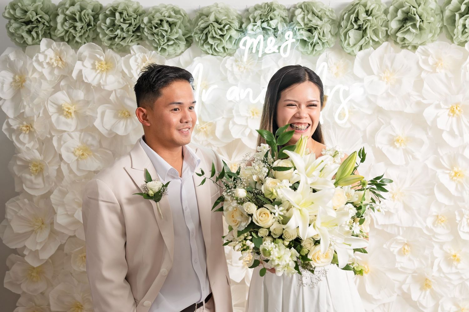 A smiling couple poses in front of a white floral wall backdrop, the bride holding a bouquet of white roses and lilies.