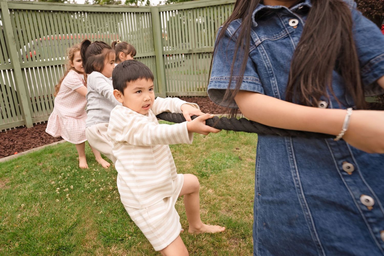 Children playing tug of war in a backyard garden, with kids laughing and pulling a rope on green grass.