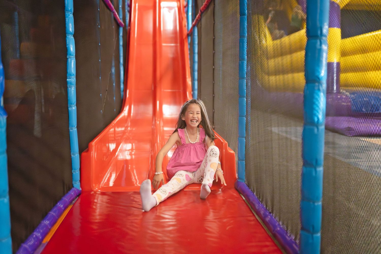 Girl smiling while sliding down colorful indoor playground slide at bounce house play center.
