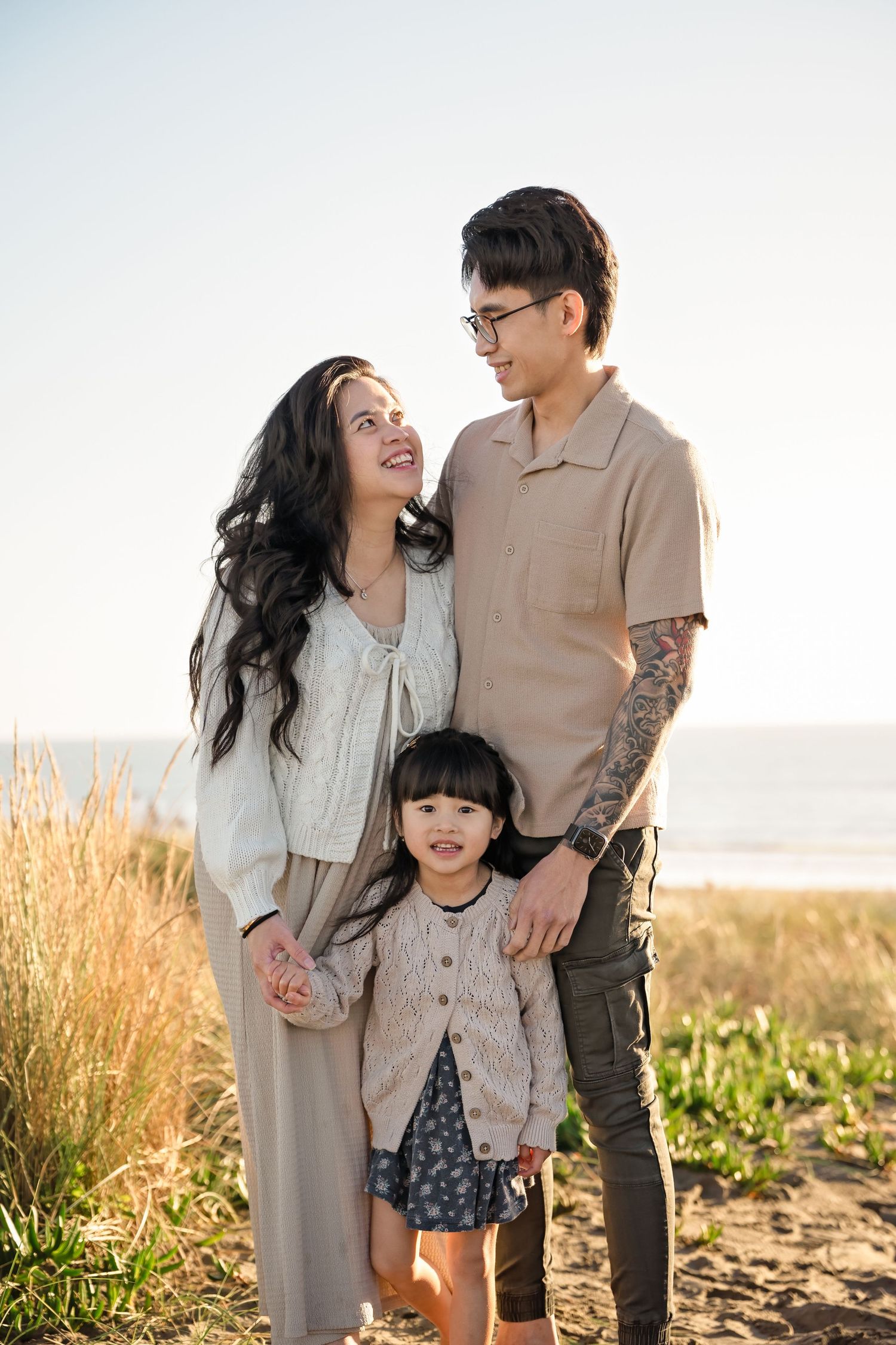 A happy Asian family of three poses outdoors near a beach, with parents smiling at each other and a young daughter between them.