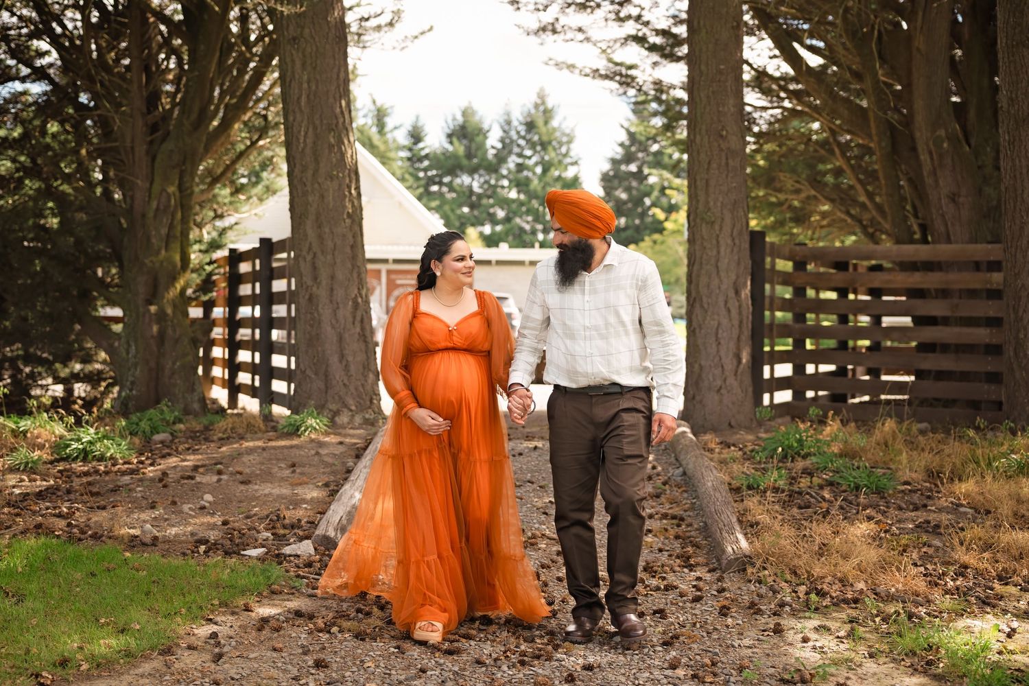 Pregnant couple in matching orange outfits holding hands on a rustic path surrounded by trees and wooden fences.