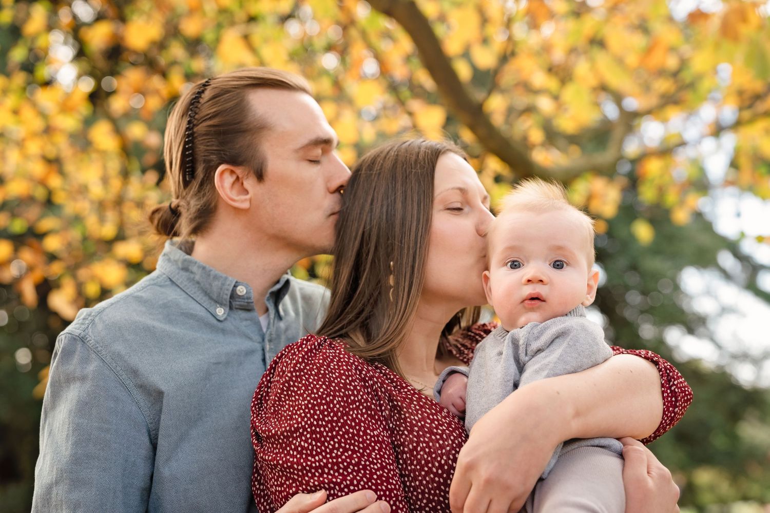 A couple kisses their baby outdoors surrounded by autumn foliage in a warm family portrait.