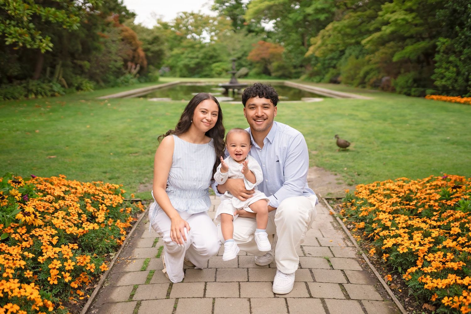 A happy family of three poses among orange flowers in a lush garden park, with parents holding their baby.