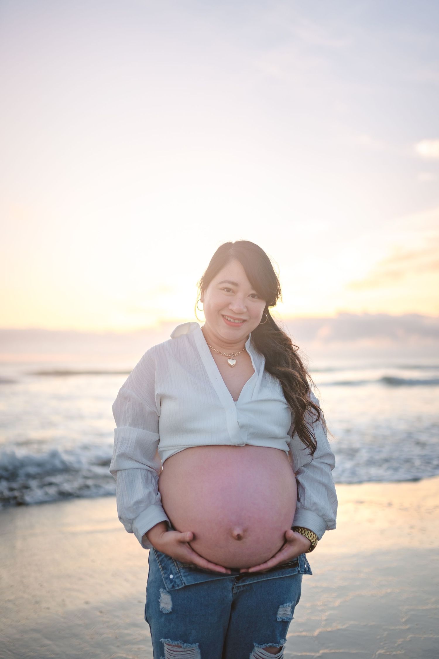 Pregnant woman in white top and jeans poses at sunset on a beach.