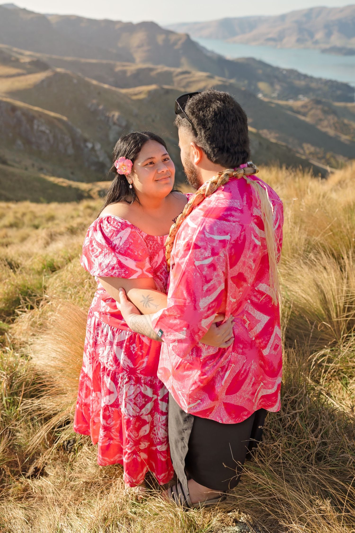 Couple in matching pink floral outfits embracing on a mountain with scenic lake views in the background.