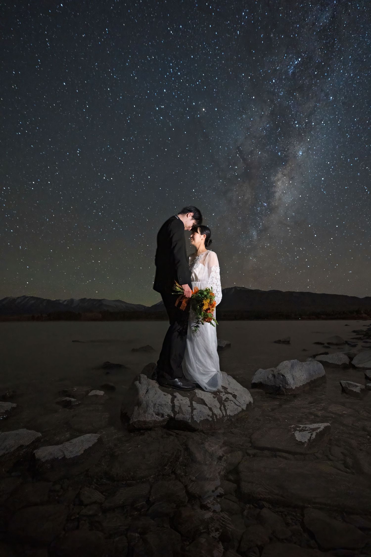 A couple sits together on coastal rocks under a starlit night sky with the Milky Way visible above.