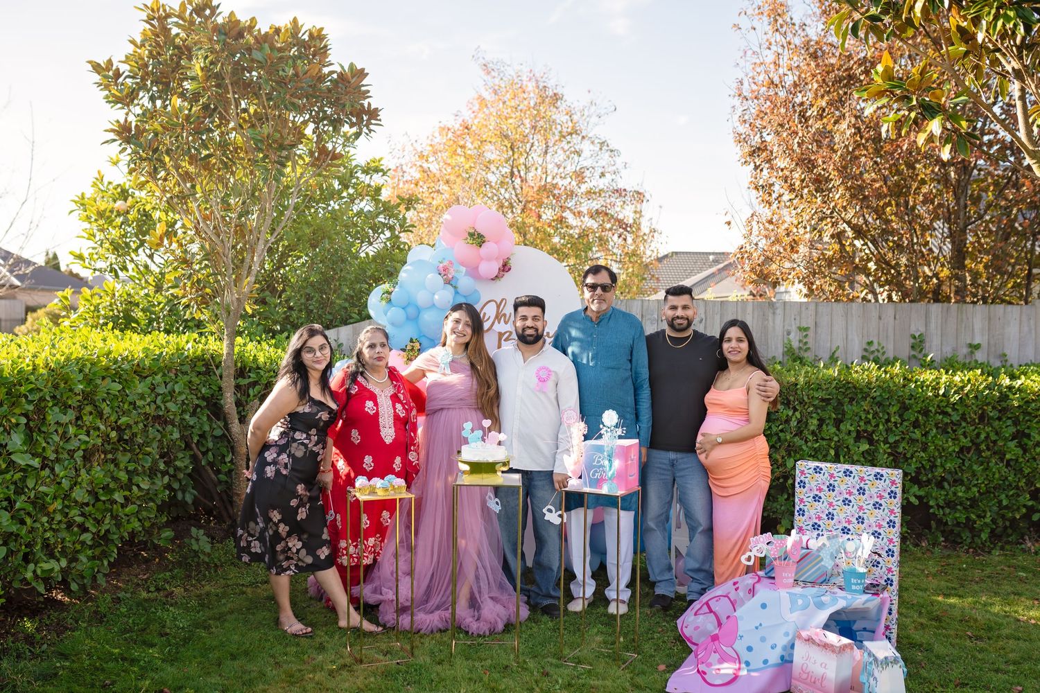 Gender reveal party celebration in backyard with balloons, presents and decorated hedge backdrop in autumn setting.