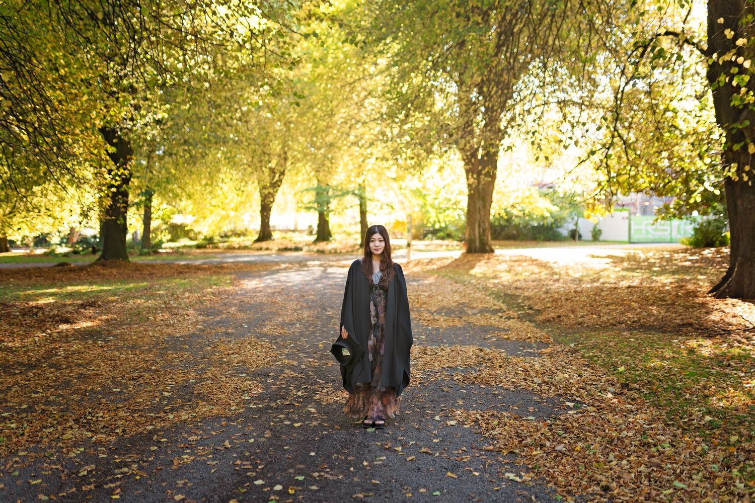 A person walks through a golden autumn park path covered in fallen leaves during sunset.
