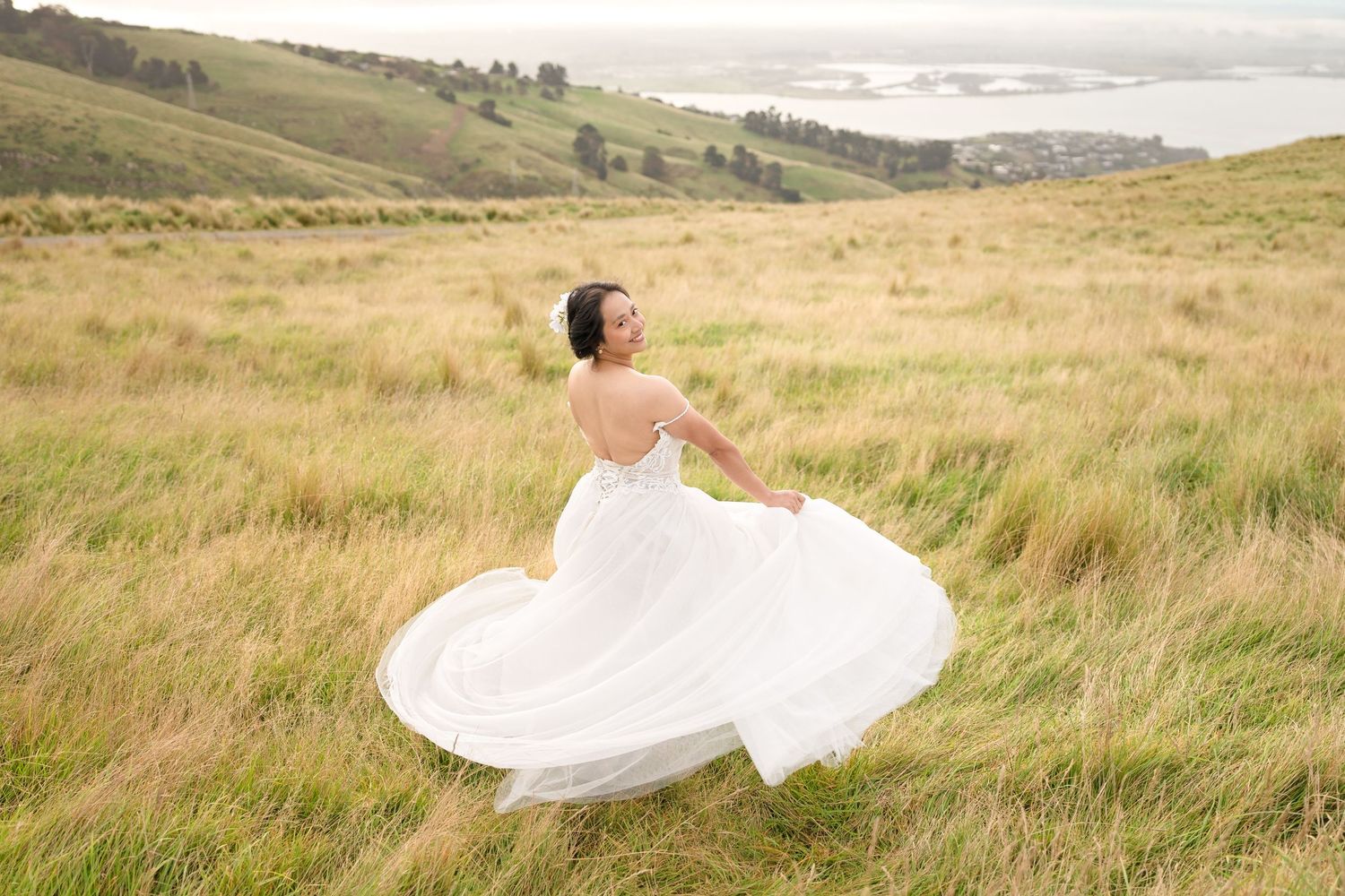Bride in white gown twirling in golden grass field with scenic coastal hills in background.