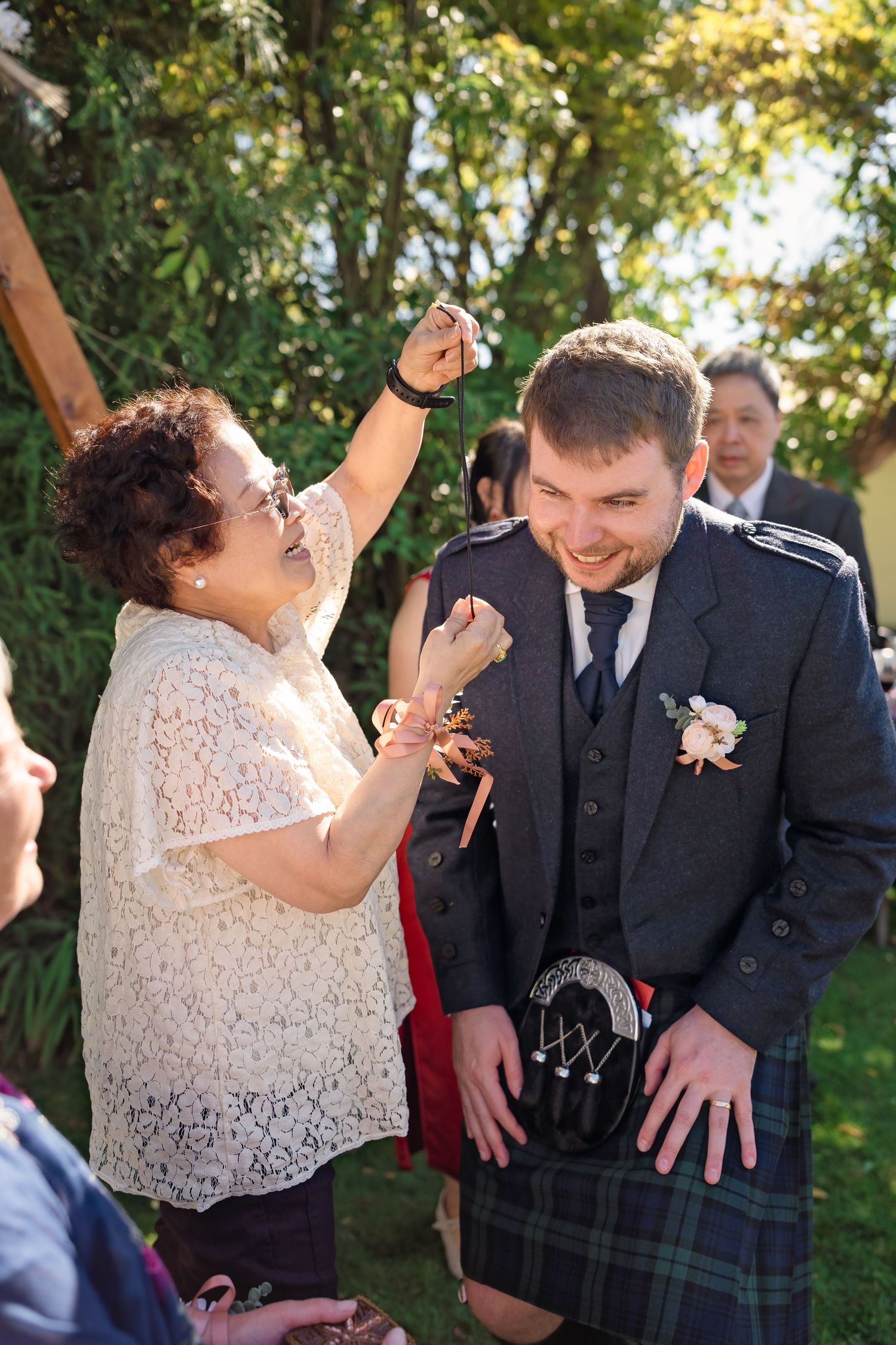 Groom in Scottish kilt and boutonniere laughs as woman playfully touches his face at outdoor wedding.