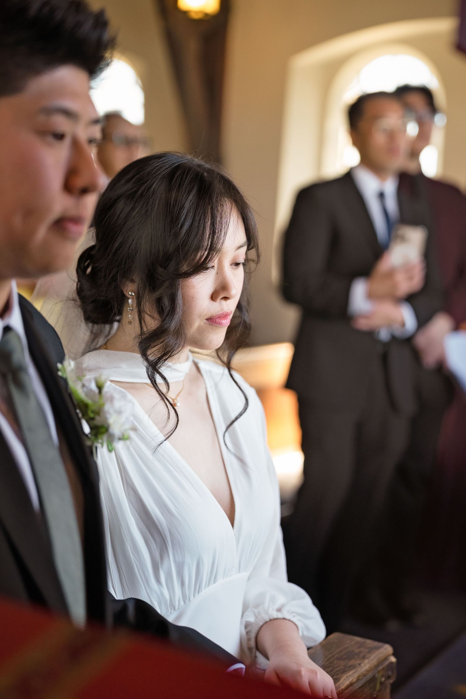 Asian bride in white dress sitting in church pew during wedding ceremony.