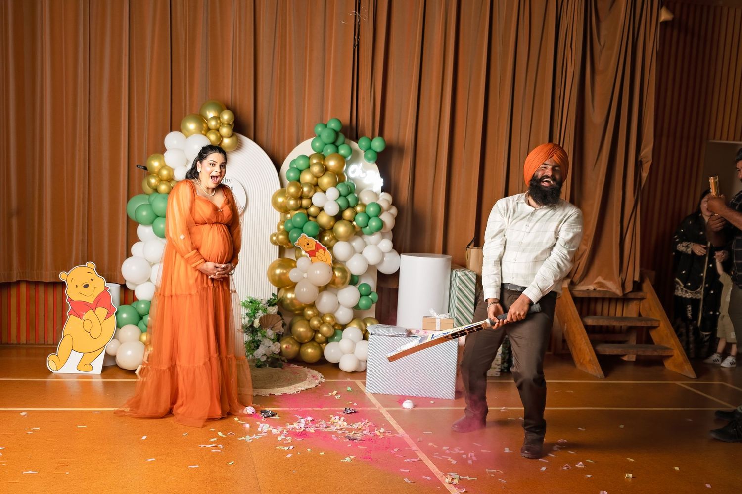 Pregnant woman in orange dress at gender reveal party as man swings bat at confetti cannon with balloon arch backdrop.